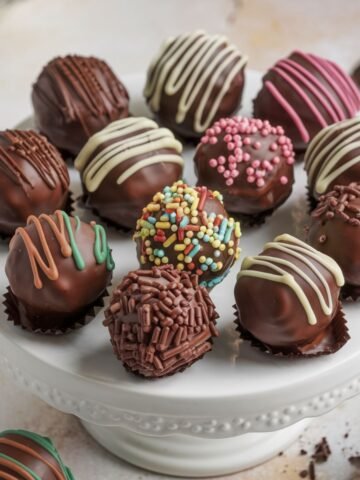 Chocolate truffle bites for Ball Cake Recipe – A variety of decorated cake balls with colorful sprinkles, chocolate drizzle, and toppings, displayed on a white cake stand.