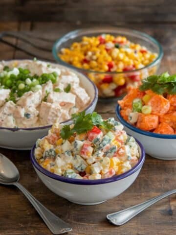 A vibrant assortment of Chicken Salad Chick recipe variations featuring creamy chicken salad, sweet potato salad, corn salad, and colorful vegetable medleys in rustic bowls on a wooden table. Perfect for fresh and flavorful meal ideas.