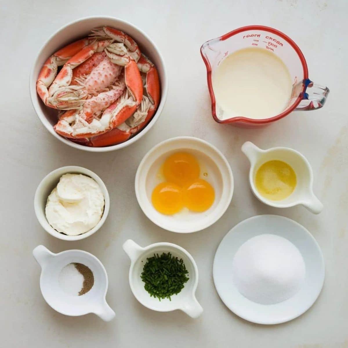 Fresh ingredients for Crab Brûlée Recipe, including crab meat, egg yolks, cream, chives, mascarpone, butter, sugar, salt, and pepper, beautifully arranged on a light background.