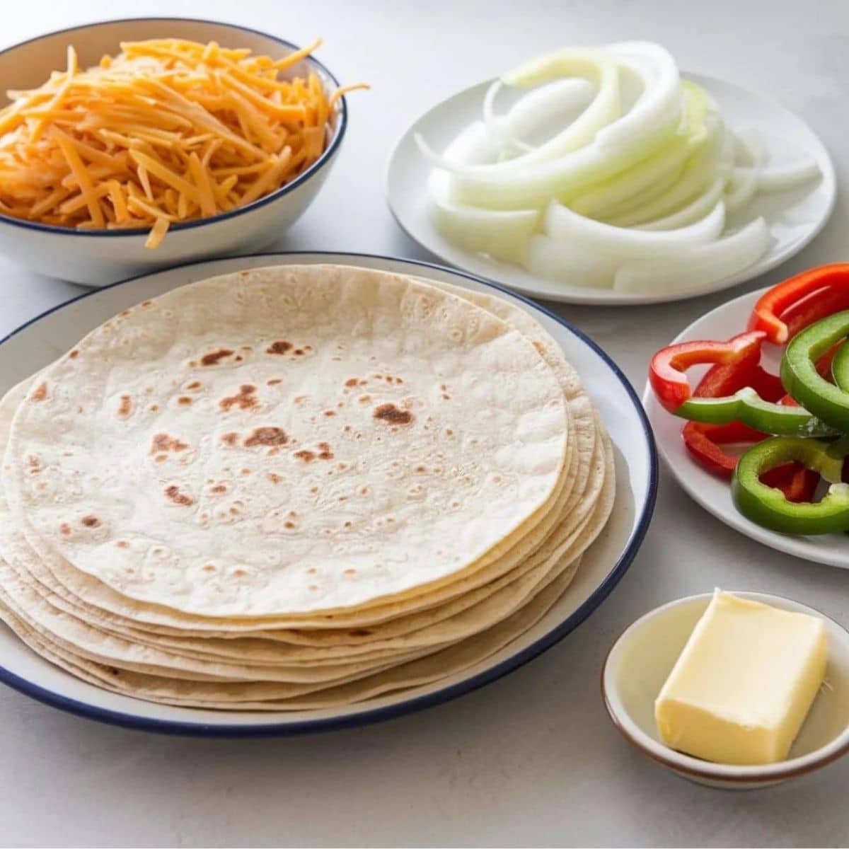 Essential ingredients for a Steak Quesadilla Recipe, featuring flour tortillas, shredded cheese, sliced onions, bell peppers, and butter, ready for assembly.