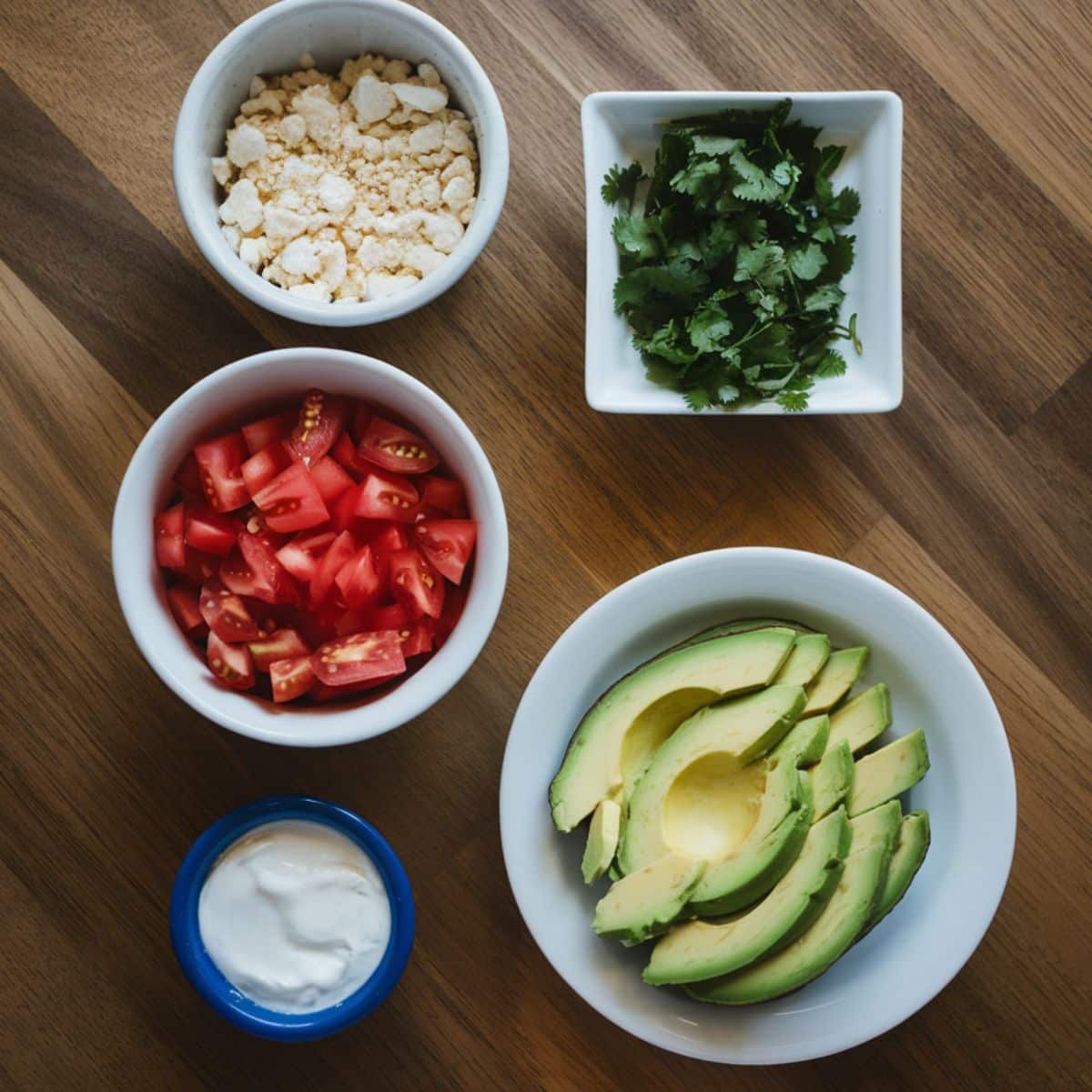 Fresh garnishes for Boulder's Enchilada Recipe including crumbled queso fresco, chopped cilantro, diced tomatoes, sliced avocado, and a bowl of sour cream, arranged on a wooden surface.