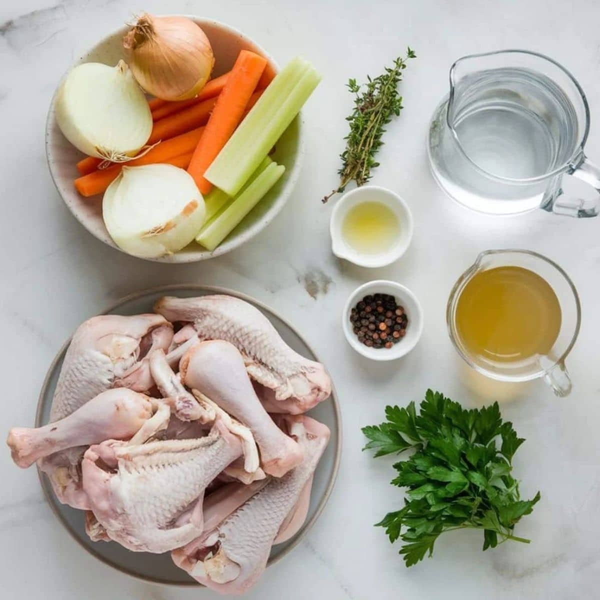 A flat-lay photo of fresh ingredients for a homemade chicken broth recipe, including raw chicken drumsticks, chopped onions, carrots, celery, parsley, thyme, black peppercorns, water, and broth, arranged on a white marble surface.