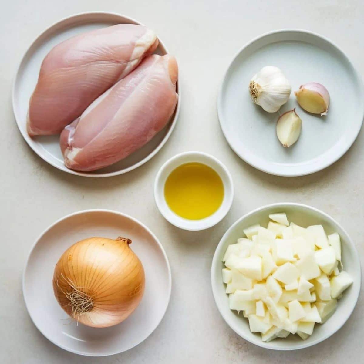 Key ingredients for chicken tacos recipe, including raw chicken breasts, garlic cloves, olive oil, onion, and diced onion, arranged on white plates.