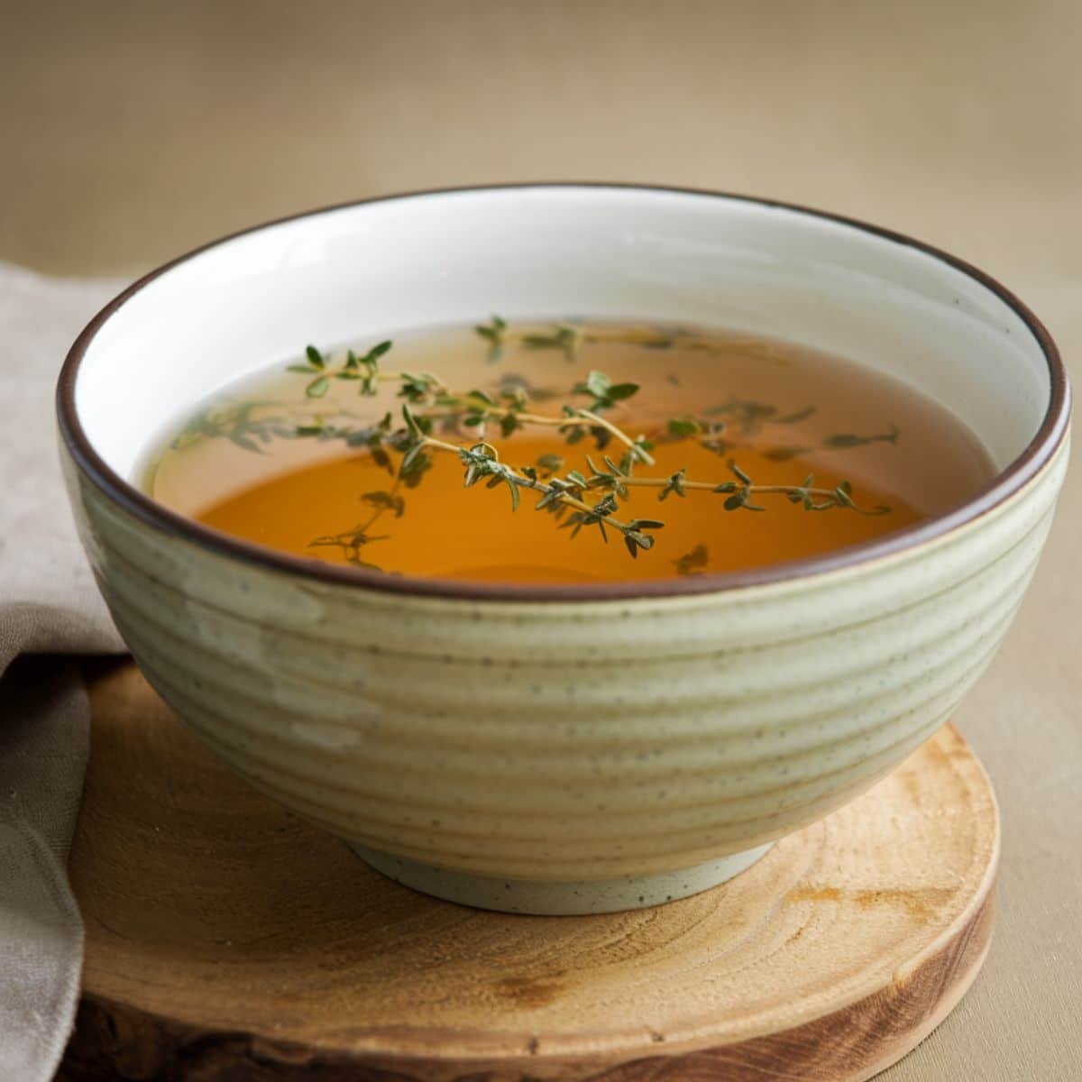 A rustic ceramic bowl filled with golden homemade chicken broth Recipe, garnished with fresh thyme sprigs, placed on a wooden cutting board. The broth has a rich, clear texture, making it perfect for soups, stews, or sipping