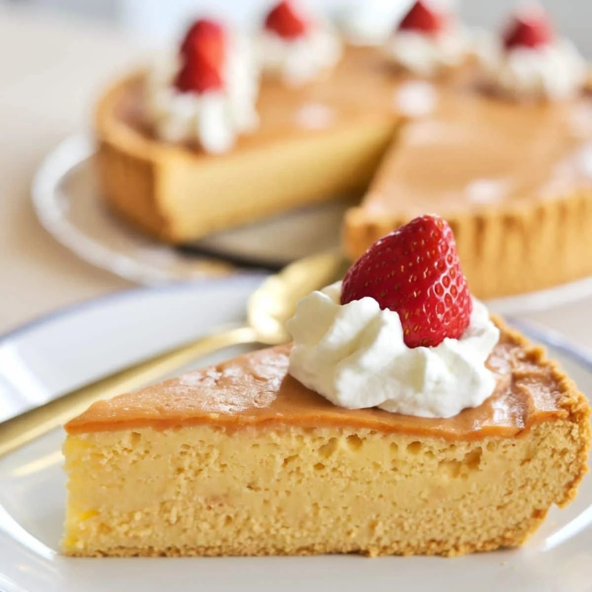 A slice of golden, creamy Impossible Pie topped with whipped cream and a fresh strawberry, served on a white plate with a full pie in the background. This easy, self-crusting Impossible Pie Recipe is a delicious and classic dessert perfect for any occasion.