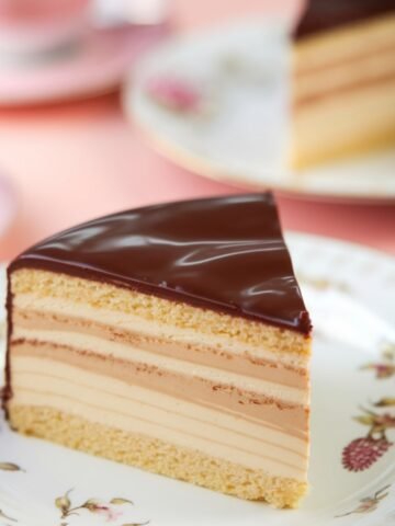 A slice of Boston Cream Cake featuring layers of fluffy vanilla sponge cake, rich custard filling, and a smooth, glossy chocolate ganache topping, served on an elegant floral plate. This classic dessert is indulgent and perfect for any occasion.