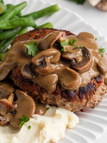 A plate of classic Salisbury steak covered in a rich and creamy mushroom gravy, served with fluffy mashed potatoes and crisp green beans. The steak is seared to perfection and topped with a savory sauce made from mushrooms, beef broth, and Worcestershire sauce. This hearty Salisbury steak recipe is the ultimate comfort food for a satisfying homemade meal.