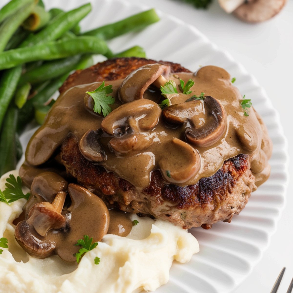 A plate of classic Salisbury steak covered in a rich and creamy mushroom gravy, served with fluffy mashed potatoes and crisp green beans. The steak is seared to perfection and topped with a savory sauce made from mushrooms, beef broth, and Worcestershire sauce. This hearty Salisbury steak recipe is the ultimate comfort food for a satisfying homemade meal.