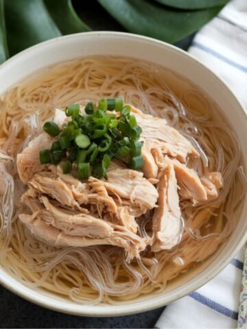 A bowl of Chicken Long Rice, a traditional Hawaiian dish, featuring tender shredded chicken, glass noodles, and a savory, ginger-infused broth. The dish is garnished with fresh green onions and served with chopsticks, making it a comforting and flavorful meal inspired by Hawaiian and Asian cuisine.