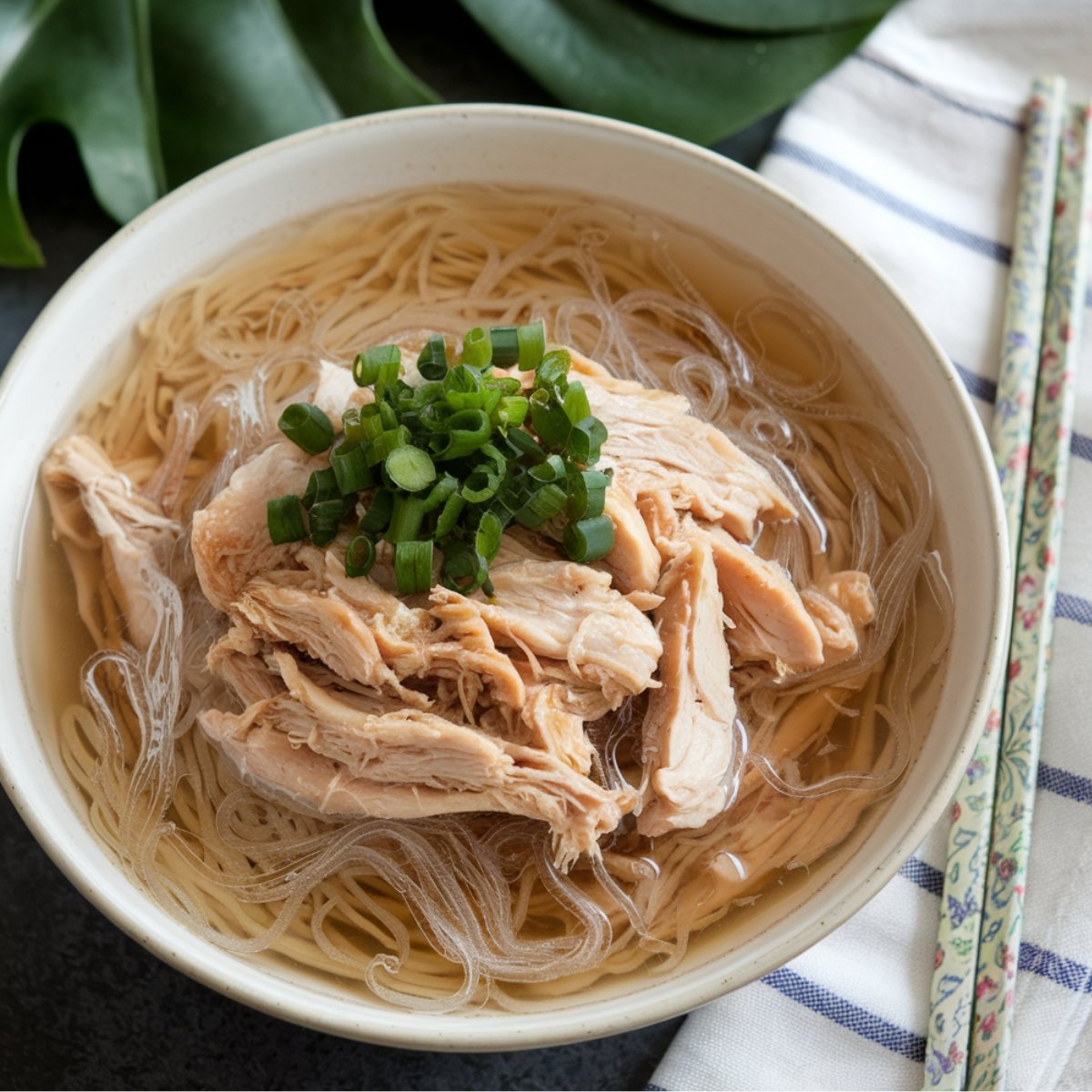 A bowl of Chicken Long Rice, a traditional Hawaiian dish, featuring tender shredded chicken, glass noodles, and a savory, ginger-infused broth. The dish is garnished with fresh green onions and served with chopsticks, making it a comforting and flavorful meal inspired by Hawaiian and Asian cuisine.