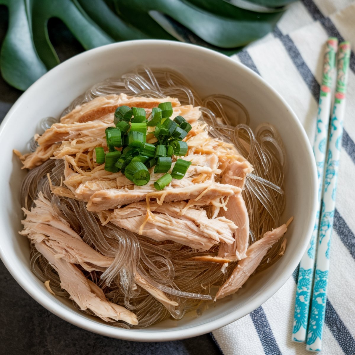 A bowl of Chicken Long Rice, a traditional Hawaiian comfort food, featuring tender shredded chicken, glass noodles, and a flavorful broth. The dish is garnished with fresh green onions and served with chopsticks, offering a warm and savory meal inspired by Hawaiian and Asian flavors.