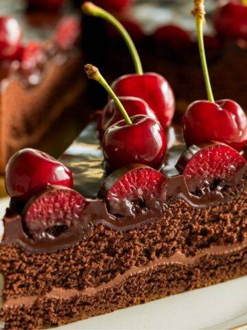 A decadent slice of Chocolate Cherry Cake Recipe featuring layers of moist chocolate sponge, rich chocolate ganache, and fresh cherries. The cake is beautifully decorated with glossy chocolate glaze and whole cherries, creating an elegant and indulgent dessert perfect for chocolate lovers.