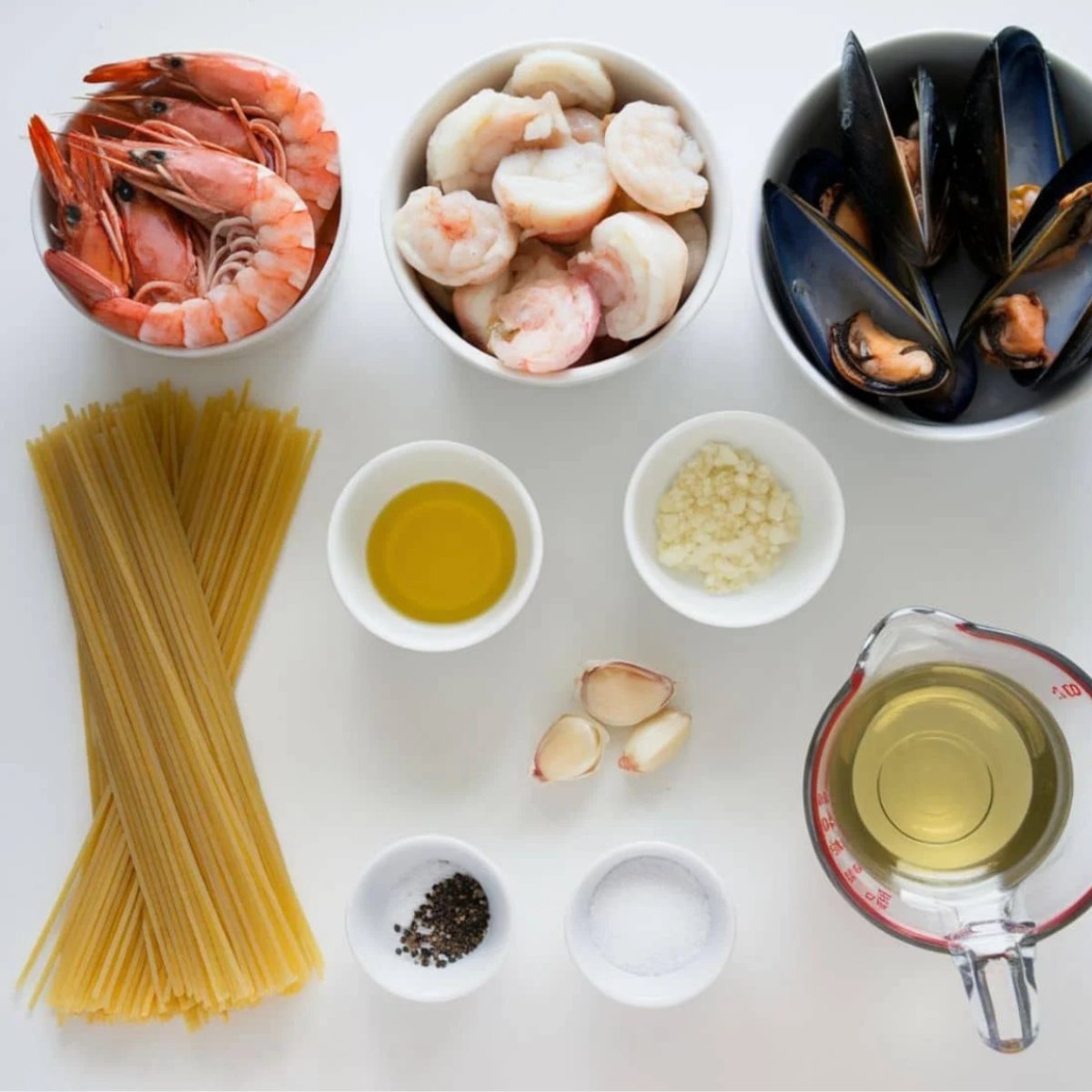 A top-down view of essential ingredients for a creamy seafood pasta recipe, including raw shrimp, mussels, spaghetti, garlic, olive oil, white wine, salt, and pepper. These fresh and flavorful ingredients create a rich and indulgent seafood pasta dish.