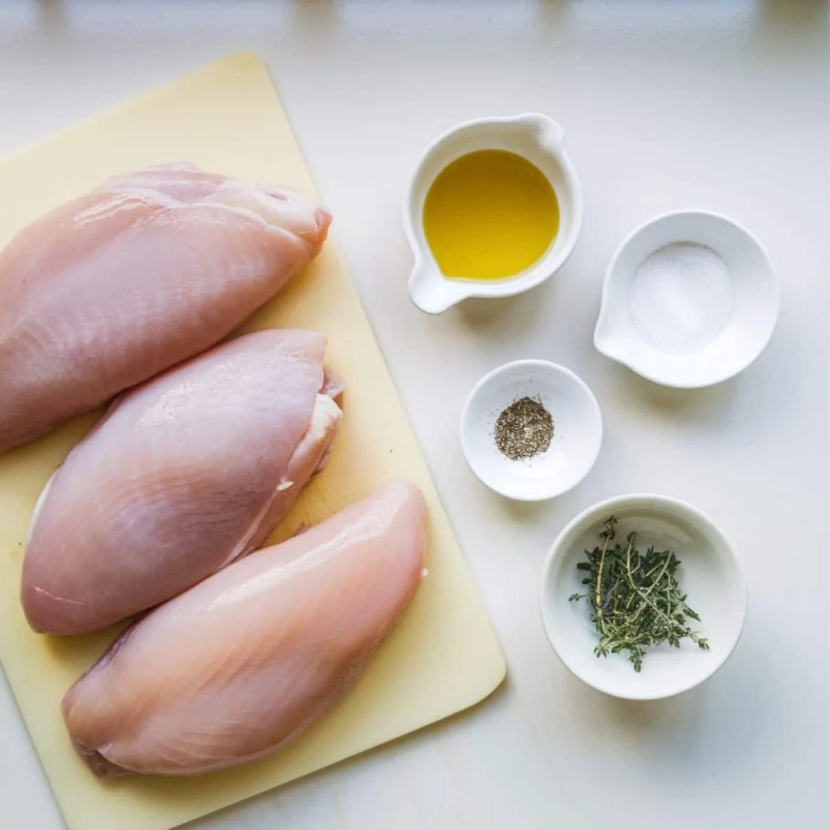 Fresh, raw chicken breasts on a cutting board, accompanied by olive oil, salt, black pepper, and fresh thyme—key ingredients for the French Onion Chicken Recipe. These simple yet flavorful seasonings help create a juicy and savory chicken base, perfectly complementing the rich caramelized onion sauce and melted cheese topping. Ideal for a comforting and delicious meal.