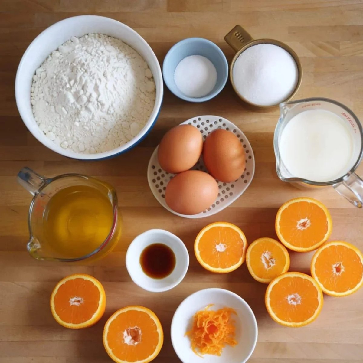 A flat lay of fresh ingredients for a Mandarin Orange Cake Recipe, including flour, sugar, eggs, milk, vanilla extract, fresh mandarin oranges, orange zest, and oil. These essential baking ingredients create a moist, citrus-flavored cake with a delightful homemade taste.