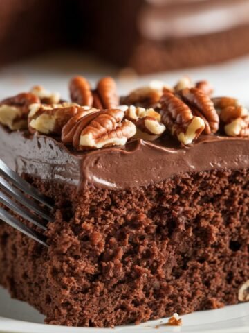 A decadent Texas Sheet Cake recipe, featuring a moist and fudgy chocolate cake topped with creamy chocolate frosting and crunchy toasted pecans. A fork digs into the rich layers, highlighting the indulgent texture of this classic Southern dessert.