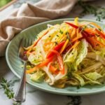 A vibrant plate of Easy Jamaican Cabbage Recipe, featuring thinly sliced cabbage, julienned carrots, and red and yellow bell peppers sautéed with fresh thyme and aromatic seasonings. The dish is served on a pastel green plate with a silver fork, set against a rustic marble countertop. A beige linen napkin and sprigs of fresh thyme add a cozy, homemade feel to this flavorful Caribbean-inspired side dish.