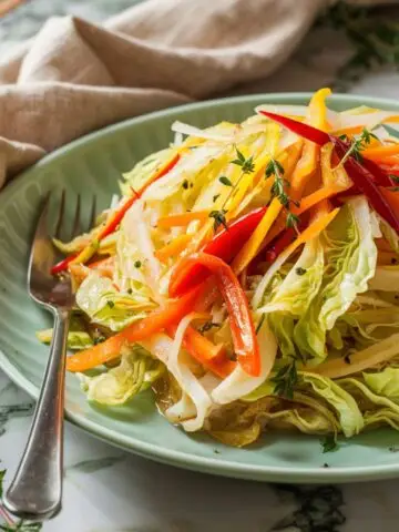 A vibrant plate of Easy Jamaican Cabbage Recipe, featuring thinly sliced cabbage, julienned carrots, and red and yellow bell peppers sautéed with fresh thyme and aromatic seasonings. The dish is served on a pastel green plate with a silver fork, set against a rustic marble countertop. A beige linen napkin and sprigs of fresh thyme add a cozy, homemade feel to this flavorful Caribbean-inspired side dish.