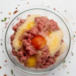 A glass bowl filled with ingredients for a Salisbury steak recipe, including ground beef, breadcrumbs, eggs, finely diced onion, and seasonings. The mixture is ready to be combined for forming tender and flavorful Salisbury steak patties.