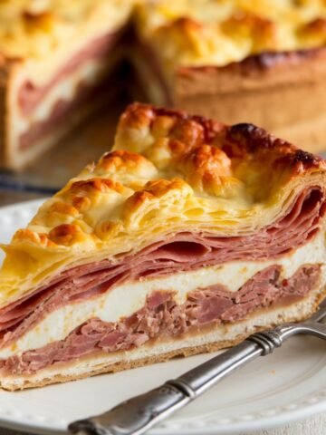 A slice of Pizza Rustica Recipe, an Italian Easter Pie, featuring layers of cured meats, ricotta cheese, and flaky golden crust, served on a white plate with a fork. A traditional savory pie perfect for Easter and special occasions.