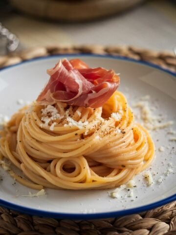 A delicious Traditional Carbonara Recipe featuring al dente spaghetti tossed in a creamy Pecorino Romano and black pepper sauce, elegantly topped with thinly sliced prosciutto. Served on a rustic white plate with a blue rim, this classic Italian dish showcases authentic flavors and textures, perfect for a comforting meal.