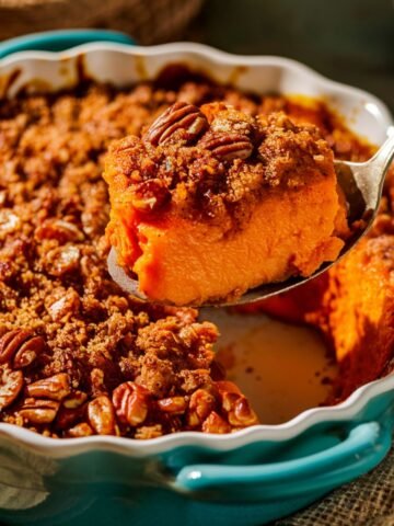 A freshly baked Ruth's Chris Sweet Potato Casserole Recipe served in a blue scalloped baking dish. A spoon lifts a creamy, smooth portion of sweet potato filling topped with a golden-brown pecan crumble. The crunchy topping is packed with caramelized pecans and brown sugar, perfectly complementing the soft, velvety sweet potato base. Warm natural lighting highlights the rich orange hue and crispy texture, creating an inviting, home-cooked feel.