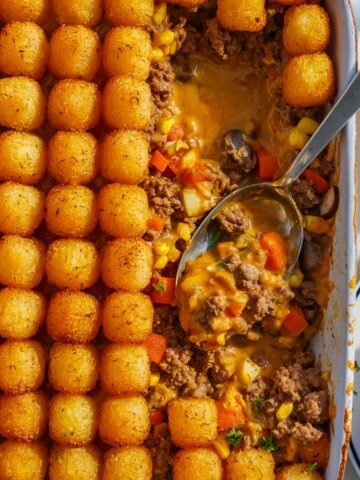 A close-up of a freshly baked Tim Walz Hot Dish Recipe in a white casserole dish. The dish features perfectly arranged, crispy golden tater tots on top of a rich, savory filling made with ground beef, diced carrots, corn, red bell peppers, mushrooms, and a creamy, cheesy sauce. A silver serving spoon scoops out a portion, revealing the hearty layers beneath. In the background, a white plate with a serving of the dish is garnished with fresh parsley, adding a touch of color. The warm lighting highlights the textures, making this classic Minnesota comfort food look extra inviting.