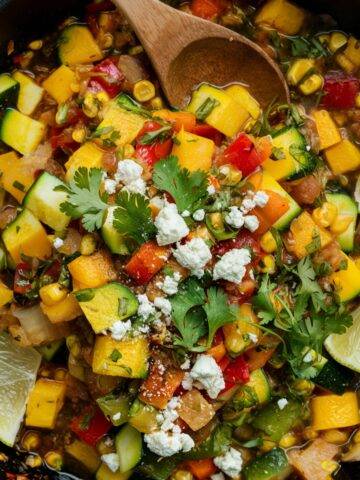 A vibrant and flavorful Calabacitas recipe in a cast-iron skillet, featuring sautéed zucchini, corn, red bell peppers, and tomatoes, garnished with fresh cilantro, crumbled queso fresco, and lime wedges. This traditional Mexican zucchini dish is a colorful and healthy vegetarian meal, perfect as a side or main course.