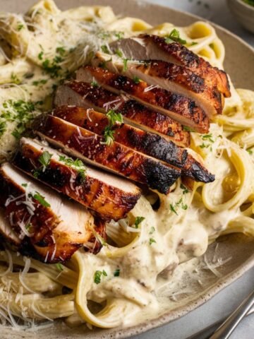 A homemade plate of Blackened Chicken Alfredo featuring creamy fettuccine pasta coated in a rich, smooth Alfredo sauce. On top, there are thick slices of blackened chicken breast, perfectly seared to a deep golden brown with a slightly charred crust, revealing a juicy, tender interior.