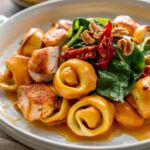 A homemade Marry Me Chicken Tortellini dish with cheese-filled tortellini coated in a creamy golden-orange sauce, topped with seared chicken, fresh spinach, sun-dried tomatoes, and toasted walnuts on a white ceramic plate.