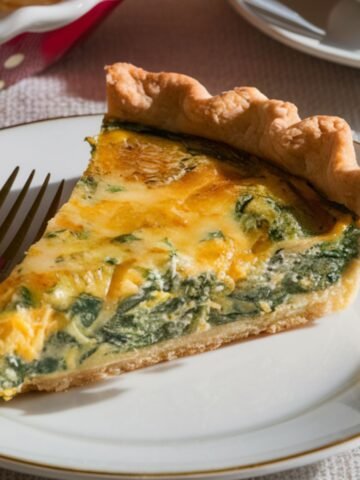 A homemade slice of spinach artichoke quiche with a golden crust, creamy filling, and vibrant spinach, served on a white and gold-rimmed plate with a fork.