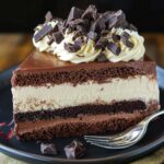 A slice of triple chocolate mousse cake with layered chocolate mousse and ganache, topped with whipped cream and dark chocolate chunks on a black plate.