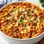 A homemade Hamburger Helper lasagna in a round white dish, topped with melted mozzarella, browned ground beef, and a sprinkle of fresh parsley. Curly lasagna noodles peek through a rich tomato meat sauce, with a blue-striped towel in the background on a white countertop.