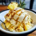 A close-up of homemade pineapple ice cream topped with caramel drizzle and fresh pineapple slices, resting on a bed of diced pineapple chunks. Soft natural light highlights the creamy texture and shiny caramel, with a spoon on the side.