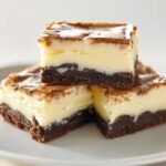 Three homemade vanilla cheesecake brownies stacked on a white plate, with a fudgy chocolate base, creamy vanilla layer, and a golden, slightly cracked top crust, lit softly to highlight the rich, real textures.