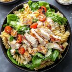 Homemade chicken Caesar pasta salad in a black bowl, with rotini pasta, fresh spinach, halved cherry tomatoes, and grilled chicken breast slices. Topped with grated Parmesan cheese and a light sprinkle of black pepper, showcasing a balance of textures and freshness.