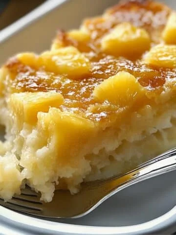 A moist, homemade pineapple dump cake slice on a white plate, topped with caramelized pineapple chunks and a glossy, golden-brown glaze, with a fork cutting into its fluffy, dense texture.