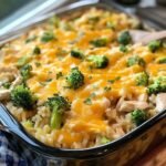 Homemade cheesy chicken broccoli rice casserole in a glass dish, topped with melted cheddar, fresh broccoli, and parsley, ready to serve with a wooden spoon.
