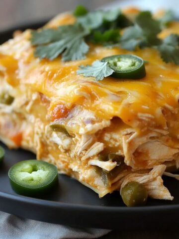 A slice of homemade chicken enchilada casserole on a black plate, topped with melted cheddar cheese, cilantro, and jalapeño slices. Layers of shredded chicken, tortillas, and veggies are visible, with extra jalapeños on the plate.