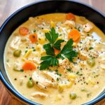 Homemade creamy chicken noodle soup with shredded chicken, egg noodles, carrots, peas, and fresh parsley in a rich, golden broth, served in a black bowl.