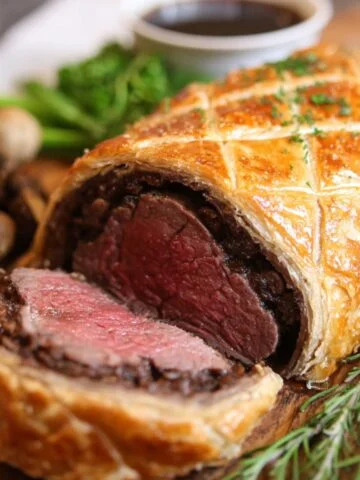 Homemade beef Wellington sliced to reveal a pink, medium-rare center wrapped in mushroom duxelles and golden, flaky puff pastry, served on a wooden board with fresh herbs and a side of gravy.
