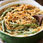 Homemade green bean casserole in a green baking dish, topped with crispy fried onions and creamy sauce, with a wooden spoon ready to serve.
