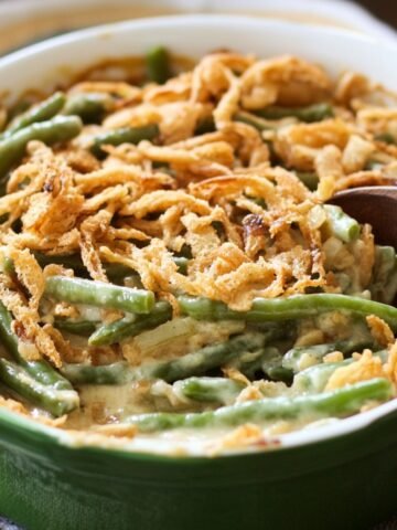 Homemade green bean casserole in a green baking dish, topped with crispy fried onions and creamy sauce, with a wooden spoon ready to serve.