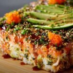 A homemade sushi bake slice on a wooden board, with layers of sushi rice and creamy seafood, topped with avocado, sesame seeds, furikake, sweet soy glaze, and bright orange masago.