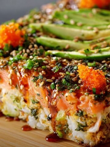 A homemade sushi bake slice on a wooden board, with layers of sushi rice and creamy seafood, topped with avocado, sesame seeds, furikake, sweet soy glaze, and bright orange masago.