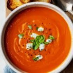 A bowl of smooth, homemade tomato soup recipe topped with basil and herbed cream, served with toasted bread on a wooden board and a spoon, with a blue napkin underneath.