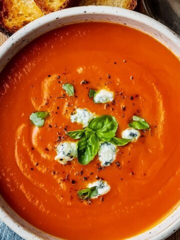 A bowl of smooth, homemade tomato soup recipe topped with basil and herbed cream, served with toasted bread on a wooden board and a spoon, with a blue napkin underneath.
