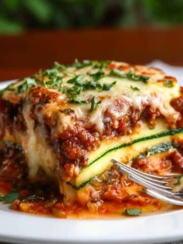 A slice of homemade zucchini lasagna with melted cheese, meat sauce, and zucchini layers on a white plate, garnished with parsley and served with a fork.