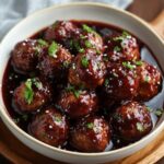 Homemade grape jelly meatballs coated in a glossy dark sauce, garnished with fresh parsley and a sprinkle of coarse salt, served in a white bowl.