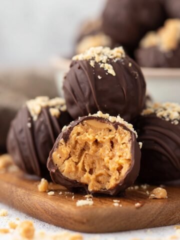 Bite-sized, homemade peanut butter balls coated in dark chocolate and drizzled with peanut butter, with one ball showing a creamy, textured peanut butter center and peanut crumbles scattered on a marble surface.